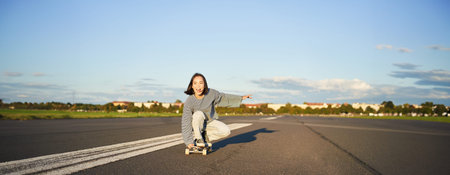 Carefree skater girl on her skateboard, riding longboard on an empty road, holding hands sideways...