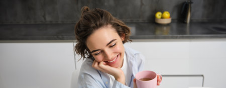 Happy mornings. Portrait of happy brunette woman, drinks cup of coffee in her kitchen and smiling, cozy and warm start of the day with cuppaの写真素材