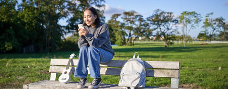 Young woman sitting in park on bench with ukulele, looking at smartphone, reading message on mobile...