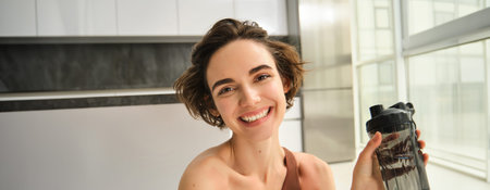 Lifestyle, sport and health concept. Portrait of happy beautiful woman, holding water bottle and smiling, wearing workout clothes at home, doing yoga exercises indoorsの写真素材