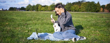 Smiling asian girl learns how to play ukulele via laptop, online video tutorials, sitting on grass in park with musical instrumentの写真素材