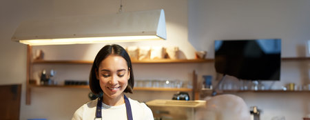 Vertical shot of friendly asian girl smiling, serving coffee, barista giving you cup of coffee,...