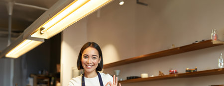 Portrait of barista girl, asian female cafe worker, shows okay, ok sign in approval, recommend smth,...