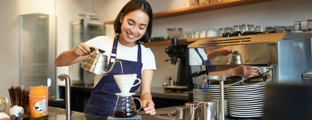 Cute brunette girl barista, cafe staff pouring water from kettle and brewing filter coffee behind counter, preparing orderの写真素材