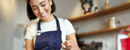 Vertical shot of brunette asian girl barista, wearing apron, making coffee, prepare order in cafe, pouring milk in cup for cappuccino latte artの写真素材