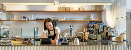 Happy smiling asian barista, girl behind counter, working with POS terminal and brewing filter kit, making coffee in cafeの写真素材