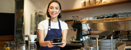 Portrait of smiling young woman providing customer service in cafe, holding cup of coffee, giving order to client, wearing apron uniformの写真素材