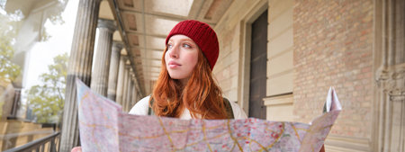 Smiling young redhead woman in red hat, looks at paper map to look for tourist attraction. Tourism...