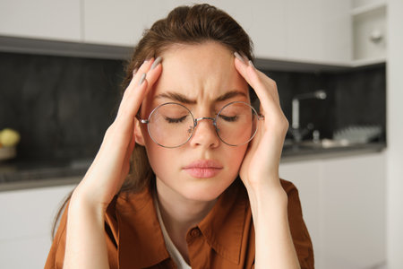 Portrait of tired young working woman in glasses, holds hands on head, suffers painful migraine, has pain in temples, sitting with headache in kitchenの写真素材