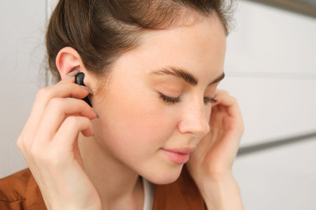 Close up portrait of tender, smiling and cute woman, puts on black wireless earphones in her ears,...