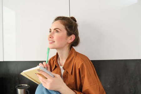 Portrait of young beautiful woman in the kitchen, looking aside and thinking, holding planner,...