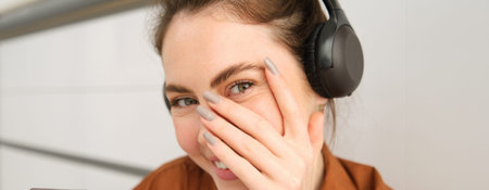 Cute smiling girl, blushes, hides face behind hand and laughing, wears wireless headphones