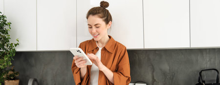 Image of beautiful young female model, posing with smartphone in the kitchen, looking at mobile...