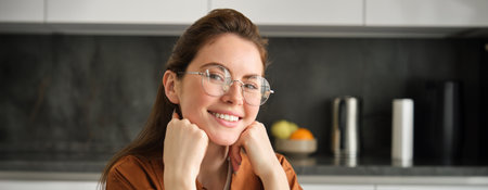 Close up portrait of young woman in her 20s, sitting in kitchen, wearing glasses, smiling and looking happy at cameraの写真素材