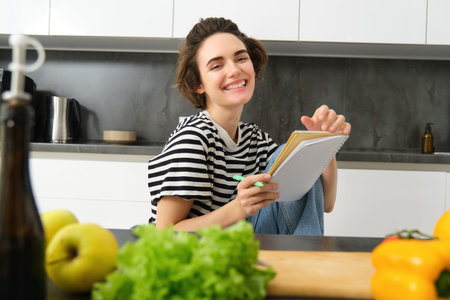 Portrait of young woman cooking, writing notes, grocery list in notebook, creating list of meals to cook through meal, sitting in kitchen near vegetables and chopping boardの写真素材