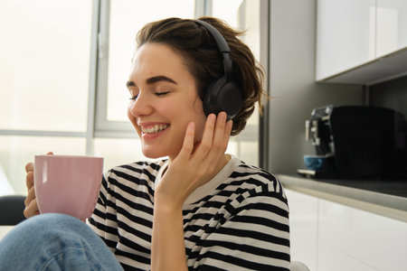 Lifestyle and music concept. Happy smiling woman with cup of tea, sits in kitchen and touches headphones on her head, listens to music with pleased relaxed faceの写真素材