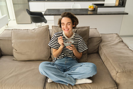 Portrait of young brunette woman eating on sofa, holding bowl and spoon, having cereals with milk...