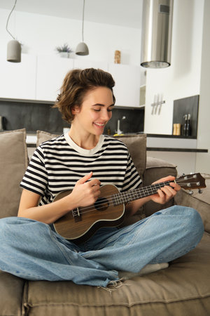 Vertical portrait of happy young woman playing ukulele, learns new musical instrument, sits on sofa at home, smiling with joyの写真素材