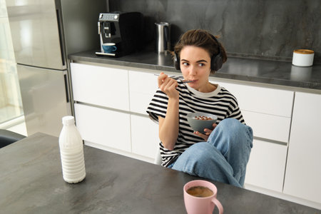 Portrait of smiling young candid woman, student eating morning cereals, having her breakfast, listening music in wireless headphones, sitting in the kitchenの写真素材