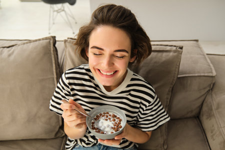 Upper angle shot of happy, cute young woman on sofa, eating bowl of cereals with milk and smiling, enjoying her breakfastの写真素材