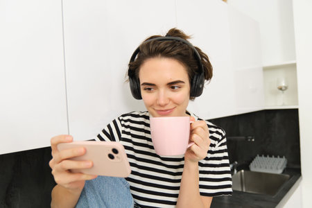 Smiling young woman watching tv series or video blog on smartphone app, drinking tea in the kitchen, looking at mobile phone with interestの写真素材
