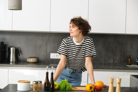 Portrait of young modern woman in the kitchen, leaning on counter with chopping board, making a salad with vegetables and dressings, cooking vegetarian mealの写真素材
