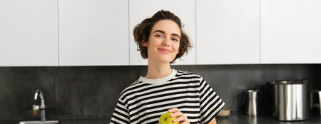 Portrait of beautiful, smiling young woman, holding an apple, eating fruit in the kitchen, looking happy. Concept of healthy diet and lifestyleの写真素材