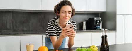 Portrait of young smiling woman, searching cooking recipe online on smartphone, standing near vegetables and chopping board, making meal, preparing salad and using social media on mobile phoneの写真素材