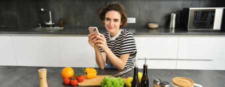 Portrait of modern young woman thinking what to cook, sitting in the kitchen with smartphone and vegetables, making a meal, healthy diet saladの写真素材