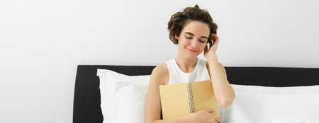 Portrait of beautiful woman in bed, sitting with her journal, writing in diary and smiling, tucking...