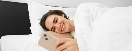 Image of woman lying in bed with white linen sheets, looking at smartphone and laughing, watching funny video on mobile phone appの写真素材