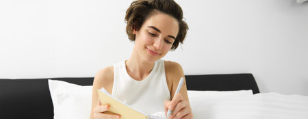 Portrait of woman sitting in bed with journal, writing in her diary, thoughts or feelings in the morning, holding notebook and smilingの写真素材