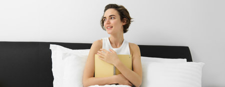 Portrait of carefree young woman writing in her journal in morning, waking up and making notes in diary, sitting in bed, smiling and looking outsideの写真素材