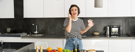 Happy beautiful woman dancing and singing while cooking in the kitchen, listening music in wireless headphones, holding smartphone, making a meal, chopping saladの写真素材