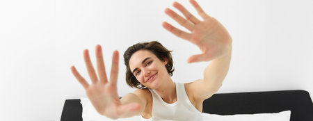 Beautiful young woman stretching her hands towards camera, waking up in morning, resting in her bed, smiling and looking happyの写真素材