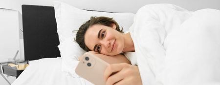 Image of female model in bed, looking at mobile phone, reading on smartphone app, smiling pleased