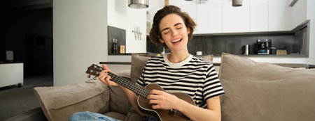 Beautiful young woman playing ukulele, singing and enjoying her musical instrument, spending time alone at home, relaxing in living roomの写真素材