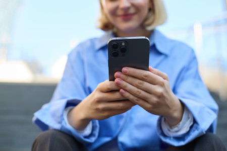 Close up portrait of young woman, holding smartphone in both hands, sitting on street with mobile phone, messaging and chatting on telephoneの写真素材