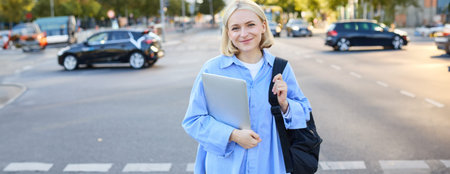 Street stylish portrait of young happy woman, blond girl with backpack and laptop, standing outdoor...
