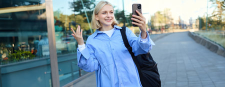 Portrait of smiling woman, lifestyle blogger recording video on smartphone, online chatting, looking at mobile camera, talking to someone online, standing on streetの写真素材