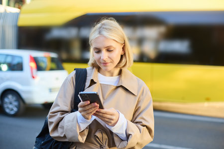 Outdoor shot of young woman with backpack, standing on street with cars passing behind her, holding mobile phone and smiling, urban lifestyle conceptの写真素材