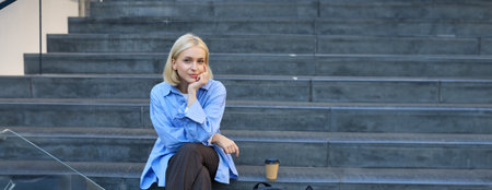 Urban style portrait of young woman, student sitting on city street stairs, with backpack, cup of coffee and laptop, looking happy and upbeat, smilingの写真素材