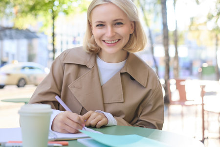 Image of young blonde woman in stylish trench, sitting in outdoor cafe, drinking coffee and working on documents, studying, doing homework in her notebook, smiling at cameraの写真素材