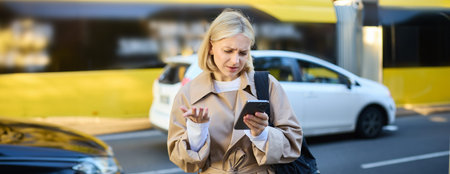 Portrait of young confused blonde woman, standing on busy street with cars behind her, looking at...