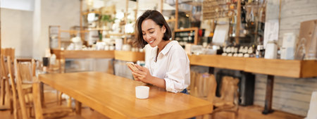 Portrait of young brunette woman, sitting with coffee and using smartphone in a cafe, chatting on mobile phoneの写真素材