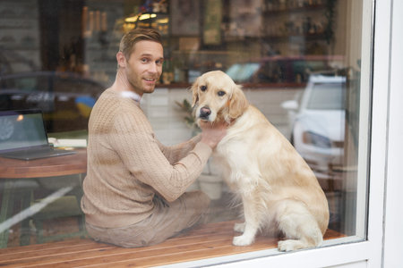 Outdoor shot of a pet-friendly cafe where man sits near the window with his fluffy golden retriever and smiles at camera. Coffee shop visitor with a dogの写真素材