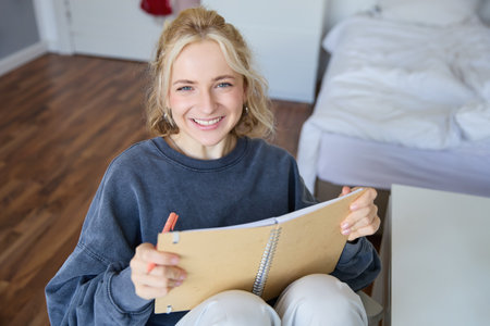 Close up portrait of beautiful smiling blond girl, holding notebook, making notes in a journal or diary, writing her thoughts, looking happy at cameraの写真素材