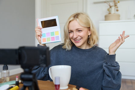 Portrait of beautiful lifestyle blogger, girl records a video on her camera for social media, shows palette of eyeshadows, does a makeup tutorial for her followers, sits in her roomの写真素材