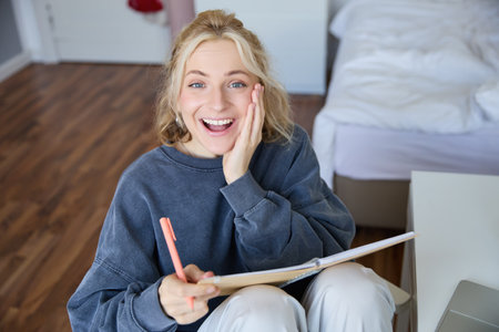 Portrait of happy, young blond woman, sitting in bedroom with notebook and pen, laughing and smiling, writing in journal, doing homework and smilingの写真素材