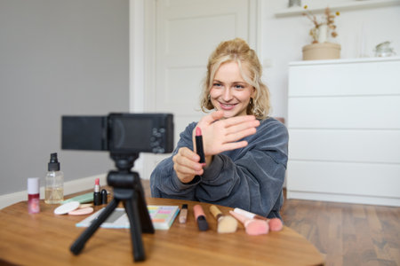 Portrait of young blond woman, teenage girl records video for her social media account, shows makeup on camera, recommends lipstick to online followers, creates content in her roomの写真素材